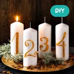 Vier weiße Adventskerzen mit goldenen Zahlen 1, 2, 3 und 4 auf einer dekorativen Unterlage, gestaltet als festliche Adventsdekoration.
